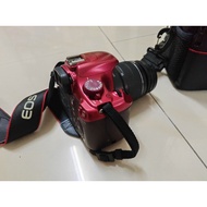 Canon EOS 1100D DSLR camera with lens, red body, black strap, on tiled floor.